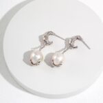 A pair of elegant earrings featuring flamingo-shaped hooks holding large natural pearls, displayed on a circular white platform with a subtle texture. Elegant pearl jewelry