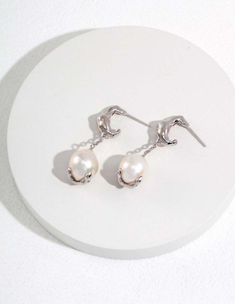 A pair of elegant earrings featuring flamingo-shaped hooks holding large natural pearls, displayed on a circular white platform with a subtle texture. Elegant pearl jewelry