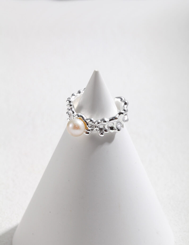 A  ring showcased on a white cone-shaped display against a light gray background. The ring features intricate metalwork with small embellishments. Elegant pearl jewelry