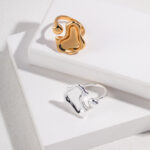 Two rings, one gold and one silver, both featuring pearl jewelry, placed on a white rectangular box with a textured background, highlighted by shadows. Elegant pearl jewelry