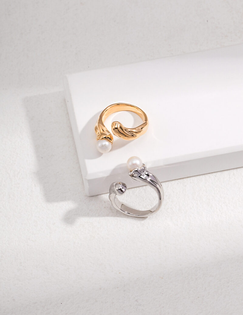 Two elegant rings displayed on a white surface; one with a natural pearl accent and a wing design, and the other with a circular clear gemstone. Elegant pearl jewelry