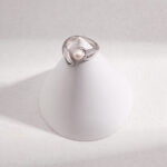 An elegant ring with a single natural pearl, displayed on a white cone-shaped stand with a textured background, highlighted by soft lighting. Elegant pearl jewelry