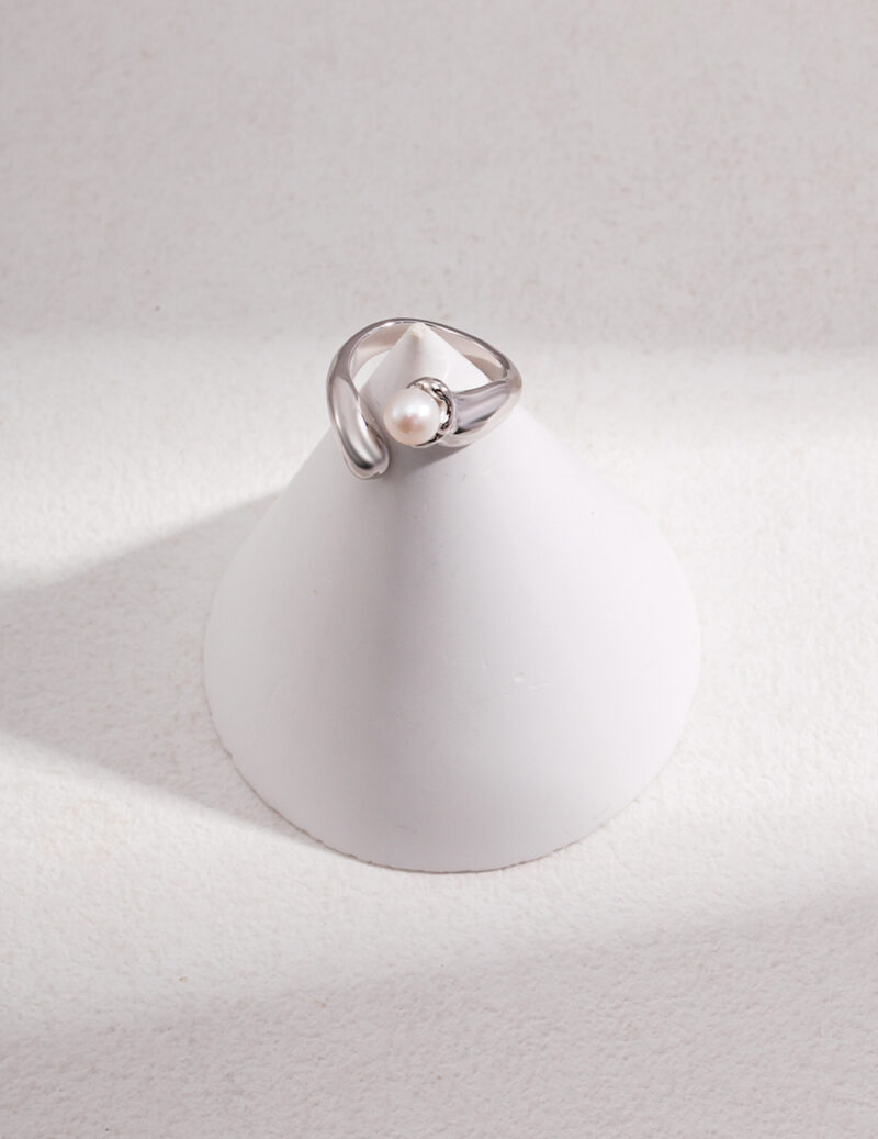 An elegant ring with a single natural pearl, displayed on a white cone-shaped stand with a textured background, highlighted by soft lighting. Elegant pearl jewelry