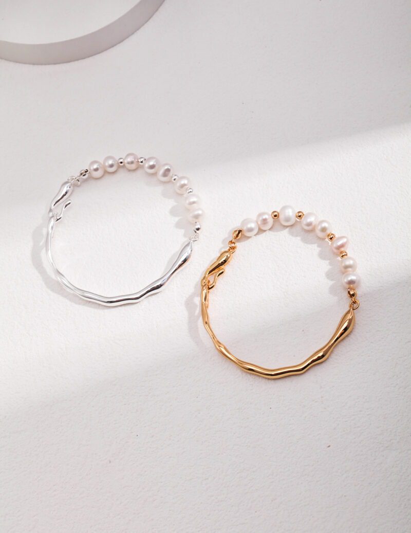 Sentence with product name: Two bracelets lying on a light surface, one with a sterling silver chain and the other with a gold chain, both featuring a wavy design. Elegant pearl jewelry