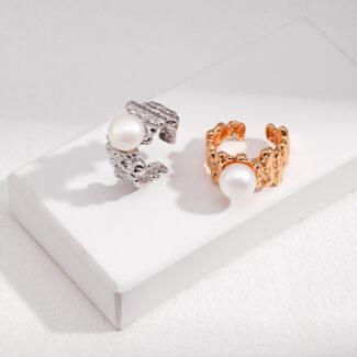 Two elegant rings with natural pearl accents displayed on a white stand against a textured background. One silver with a detailed design, and the other is gold with a link chain pattern. Elegant pearl jewelry