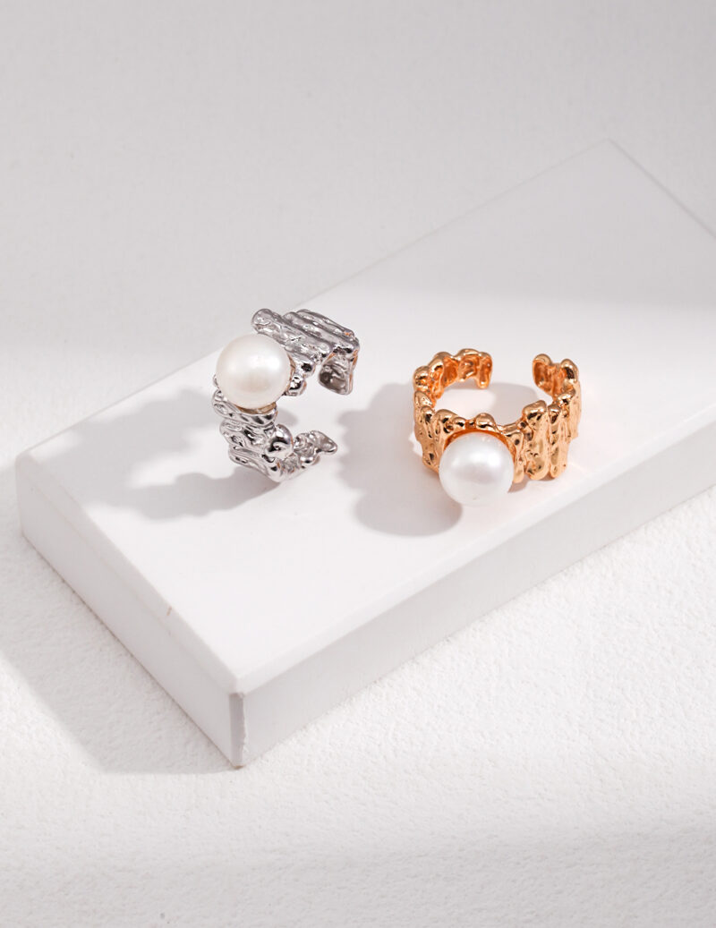 Two elegant rings with natural pearl accents displayed on a white stand against a textured background. One silver with a detailed design, and the other is gold with a link chain pattern. Elegant pearl jewelry