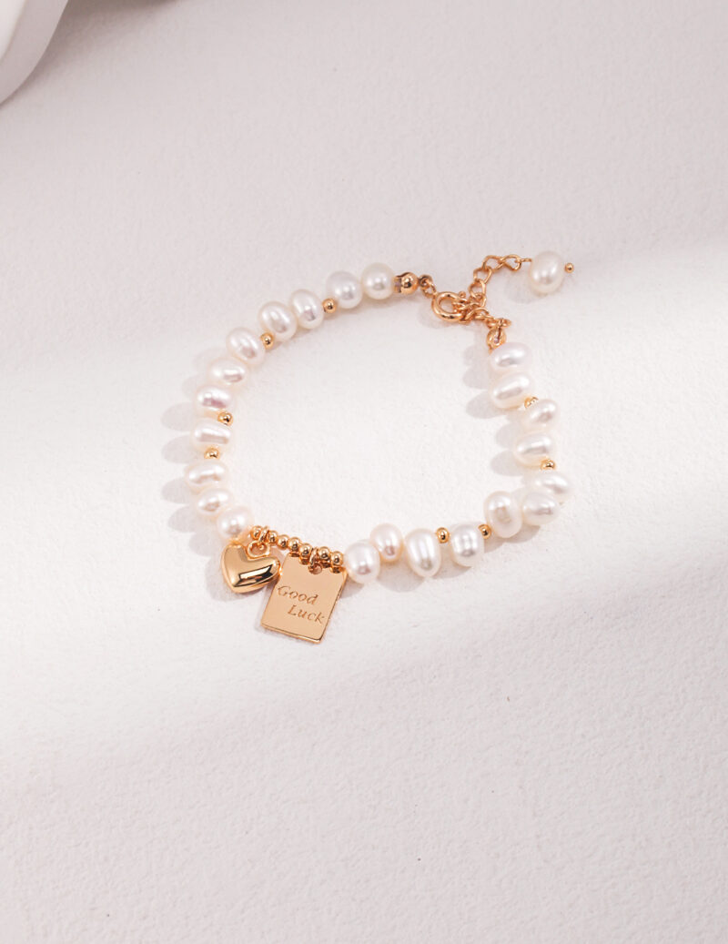 An elegant bracelet with a rose gold chain, featuring a charm that reads "good luck", laid flat on a light neutral surface. Elegant pearl jewelry