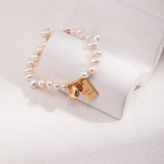 A bracelet with a gold clasp and a pendant engraved with "good luck" rests elegantly on a white stone-like jewelry display. Elegant pearl jewelry