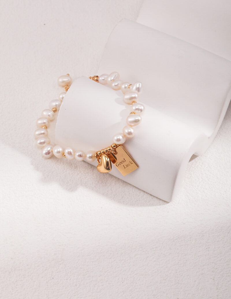 A bracelet with a gold clasp and a pendant engraved with "good luck" rests elegantly on a white stone-like jewelry display. Elegant pearl jewelry