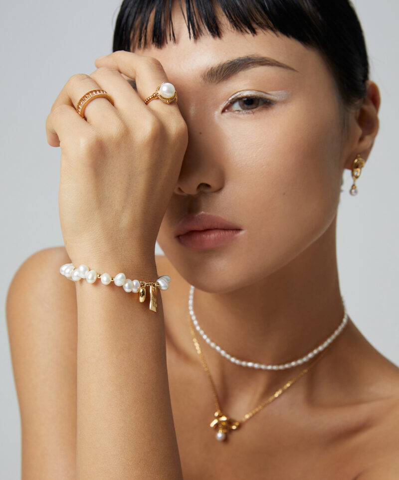 Close-up of a woman with elegant Desiree freshwater pearl jewelry, showcasing a bracelet, rings, and necklace. She has subtle makeup and a contemplative expression. Elegant pearl jewelry