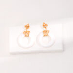 A pair of elegant with gold floral motifs and gold pearl jewelry attached to large white circular hoops, displayed on a white shelf against a white textured wall. Elegant pearl jewelry