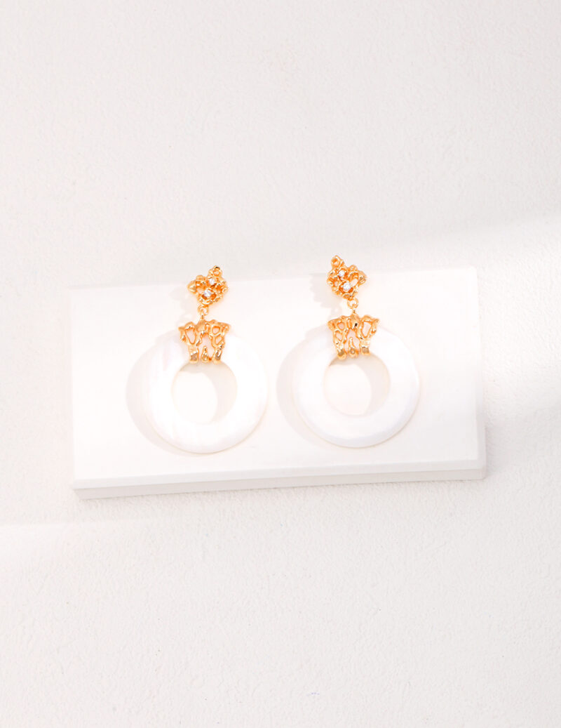 A pair of elegant with gold floral motifs and gold pearl jewelry attached to large white circular hoops, displayed on a white shelf against a white textured wall. Elegant pearl jewelry