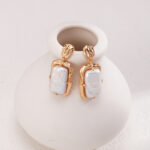 A pair of elegant earrings with rectangular opalescent stones, displayed on a small, round ceramic vase with a matte finish, positioned against a soft white background. Elegant pearl jewelry