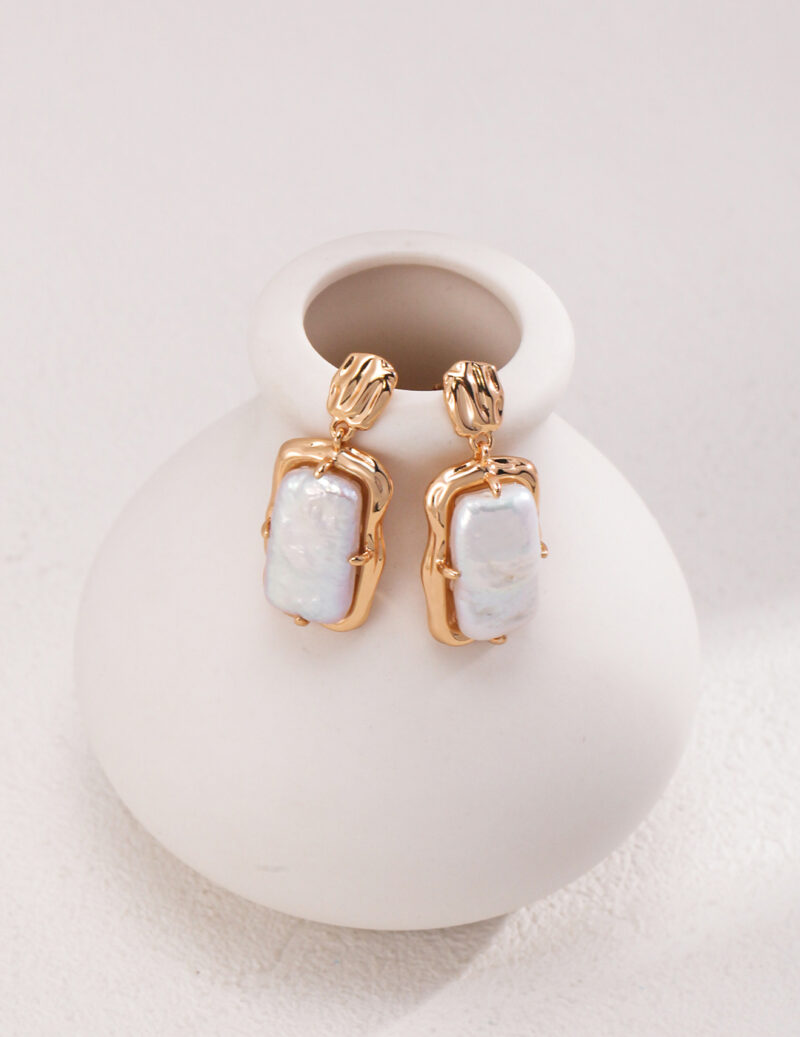 A pair of elegant earrings with rectangular opalescent stones, displayed on a small, round ceramic vase with a matte finish, positioned against a soft white background. Elegant pearl jewelry