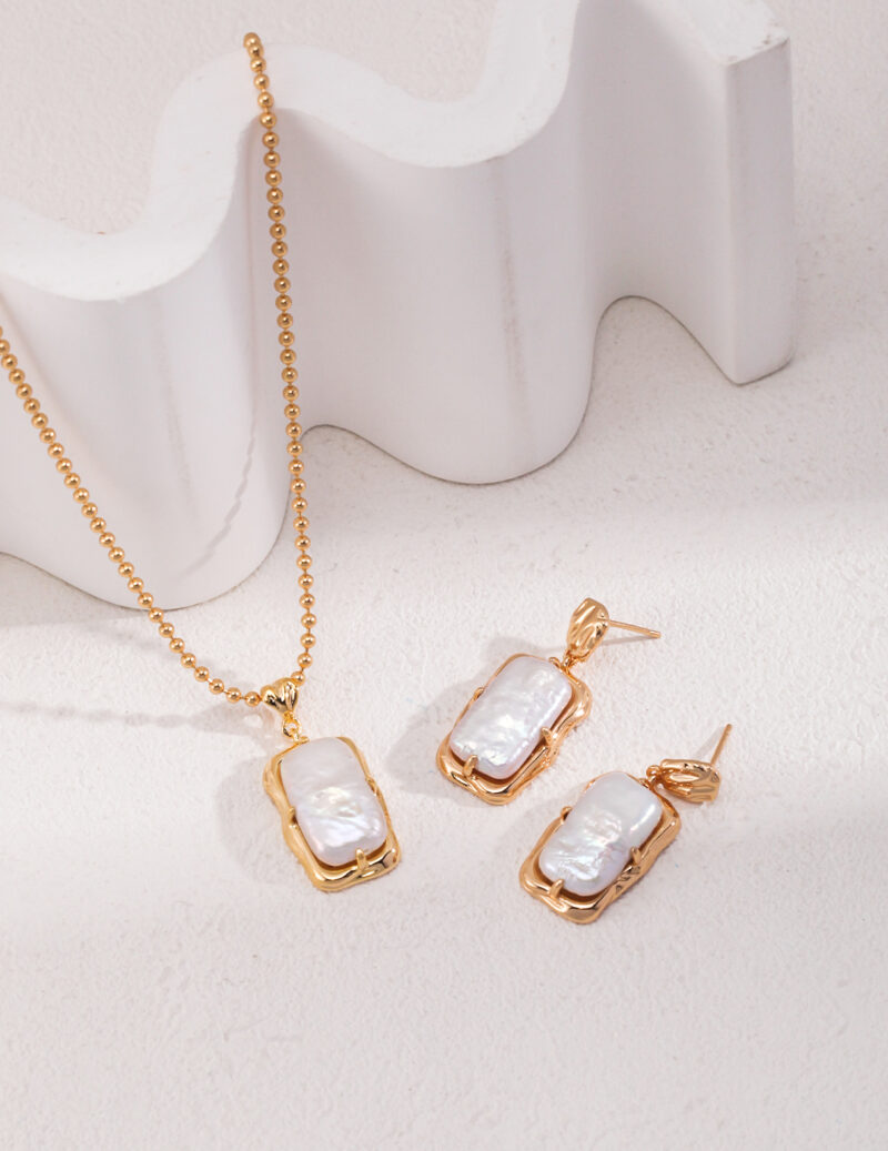 Displayed on a white surface, the Baroque Pearl Necklace collection features a gold necklace named o_1hj9givdk15n63bqllovtedd72, adorned with a rectangular white stone pendant and finished in 18k gold vermeil. The scene is complemented by a matching pair of earrings, and the background showcases a wavy white decorative element, enhancing the overall elegance. Elegant pearl jewelry