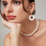 A woman with dark hair gazes thoughtfully, resting her chin on her hand. She wears lustrous white circular earrings, an o_1hkiinm2i1faqse1pmk1t5g11a13 necklace, and a matching bracelet. Her stylish outfit is adorned with pastel sequins, creating a perfect harmony with her soft makeup. Elegant pearl jewelry