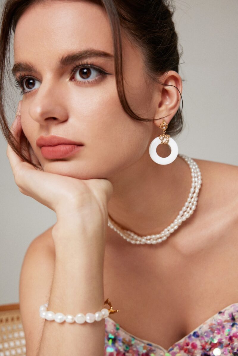 A woman with dark hair gazes thoughtfully, resting her chin on her hand. She wears lustrous white circular earrings, an o_1hkiinm2i1faqse1pmk1t5g11a13 necklace, and a matching bracelet. Her stylish outfit is adorned with pastel sequins, creating a perfect harmony with her soft makeup. Elegant pearl jewelry