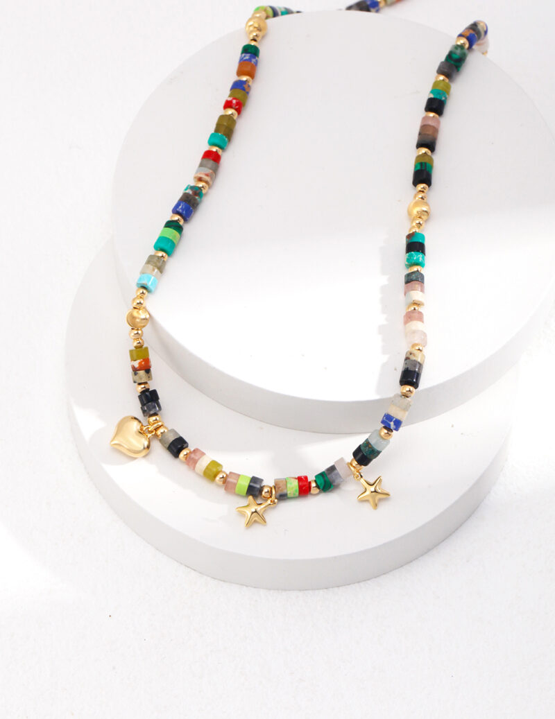 A colorful beaded necklace featuring gold star charms and a freshwater pearl, with a heart-shaped clasp, displayed on a white cylindrical stand against a white background. Elegant pearl jewelry