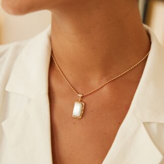 A close-up of a person wearing a white shirt showcases the o_1i3cb3lch1meb976tcg1ctm1gf89—a stunning 18k gold vermeil necklace featuring a rectangular pendant with a white stone. Partially visible, matching gold earrings with similar white stones complement the look. The ensemble, accentuated by an elegant Baroque pearl necklace, exudes a minimalistic yet refined charm. Elegant pearl jewelry