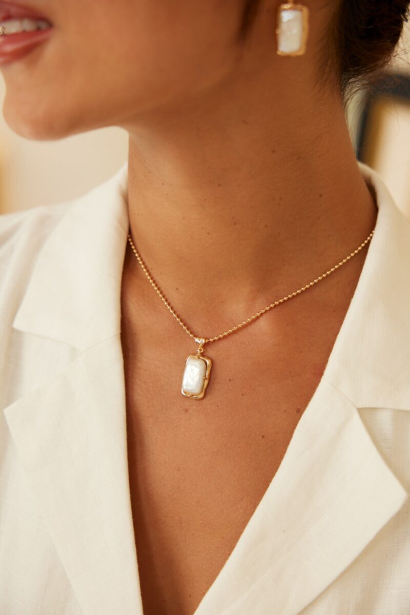 A close-up of a person wearing a white shirt showcases the o_1i3cb3lch1meb976tcg1ctm1gf89—a stunning 18k gold vermeil necklace featuring a rectangular pendant with a white stone. Partially visible, matching gold earrings with similar white stones complement the look. The ensemble, accentuated by an elegant Baroque pearl necklace, exudes a minimalistic yet refined charm. Elegant pearl jewelry