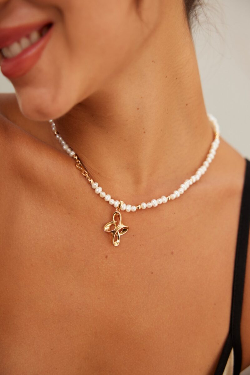 A close-up image of a woman wearing the o_1i3jqal0div4rfkpjh189algh2, a pearl necklace with an 18k gold flower pendant. The woman is smiling, and the photograph captures part of her neck and shoulder. She is dressed in a sleeveless top. Elegant pearl jewelry