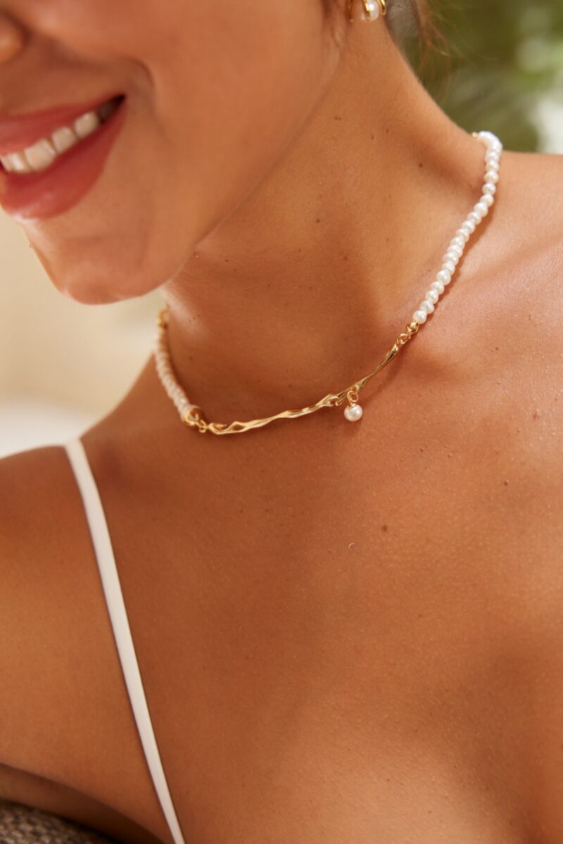 Close-up of a woman wearing the o_1i3pkk1eu161u1ivhfk81rlv16l32, featuring small white beads and an elegant twisted gold vermeil centerpiece with a small pearl hanging from it. She is smiling slightly, and the thin strap of a white top is visible on her shoulder. Elegant pearl jewelry