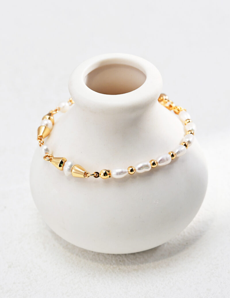 An elegant bracelet with gold accents draped over a small white ceramic vase on a light textured surface. Elegant pearl jewelry