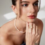 A woman with a contemplative expression rests her chin on her hand, wearing a navy dress and adorned with natural pearl jewelry, including earrings, a necklace, and a bracelet. Her tattoo is partly visible