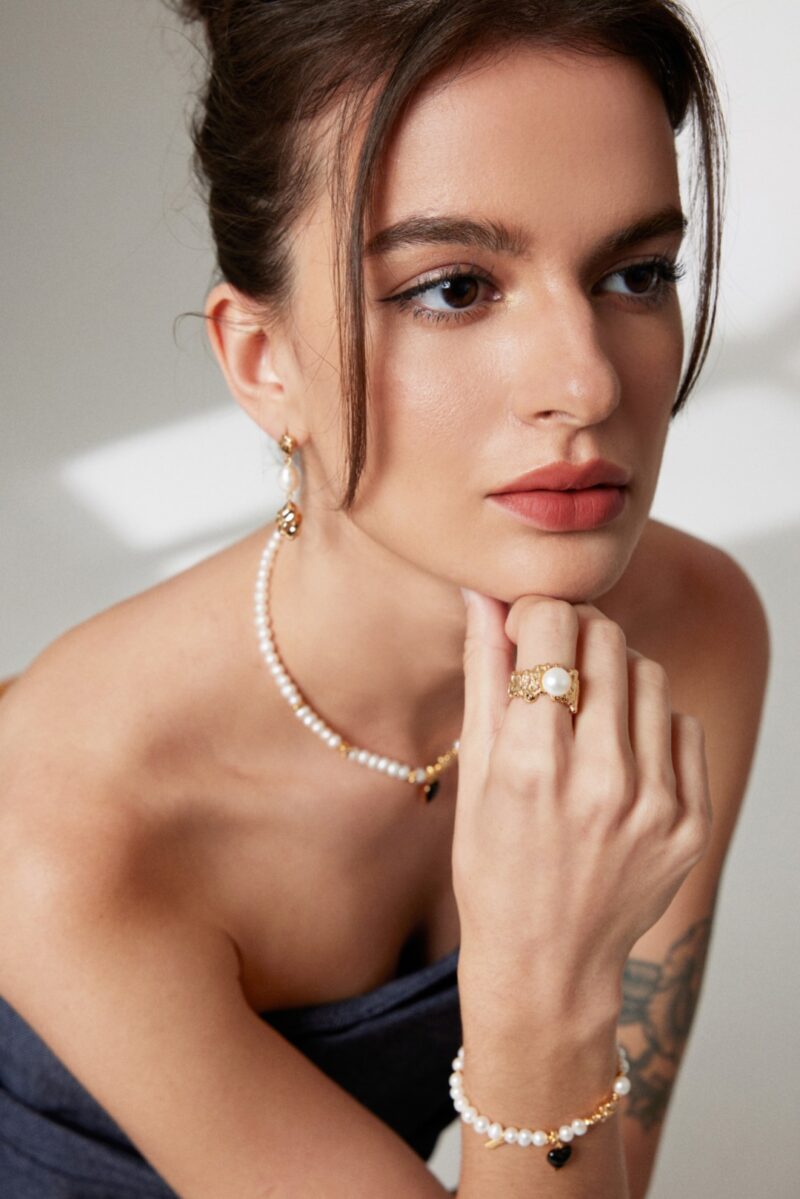 A woman with a contemplative expression rests her chin on her hand, wearing a navy dress and adorned with natural pearl jewelry, including earrings, a necklace, and a bracelet. Her tattoo is partly visible