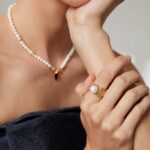 A close-up of a woman wearing jewelry, including a necklace, bracelet, and rings. She is dressed in a dark strapless top and posed with her hand touching her chin. Elegant pearl jewelry