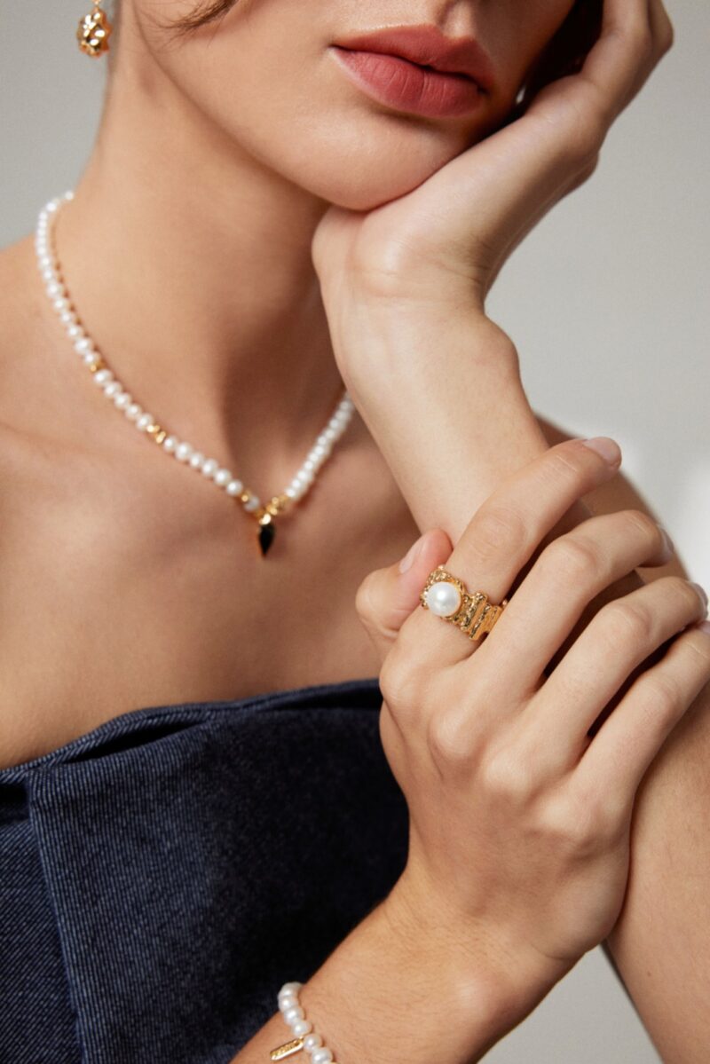 A close-up of a woman wearing jewelry, including a necklace, bracelet, and rings. She is dressed in a dark strapless top and posed with her hand touching her chin. Elegant pearl jewelry