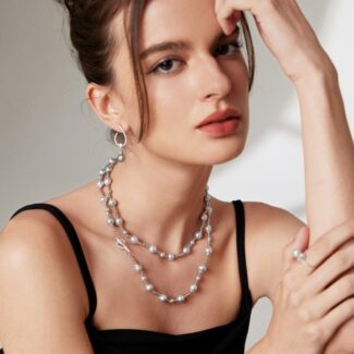 A woman with a thoughtful expression, wearing a black dress and adorned with necklace and large hoop earrings, rests her chin on her hand. Elegant pearl jewelry