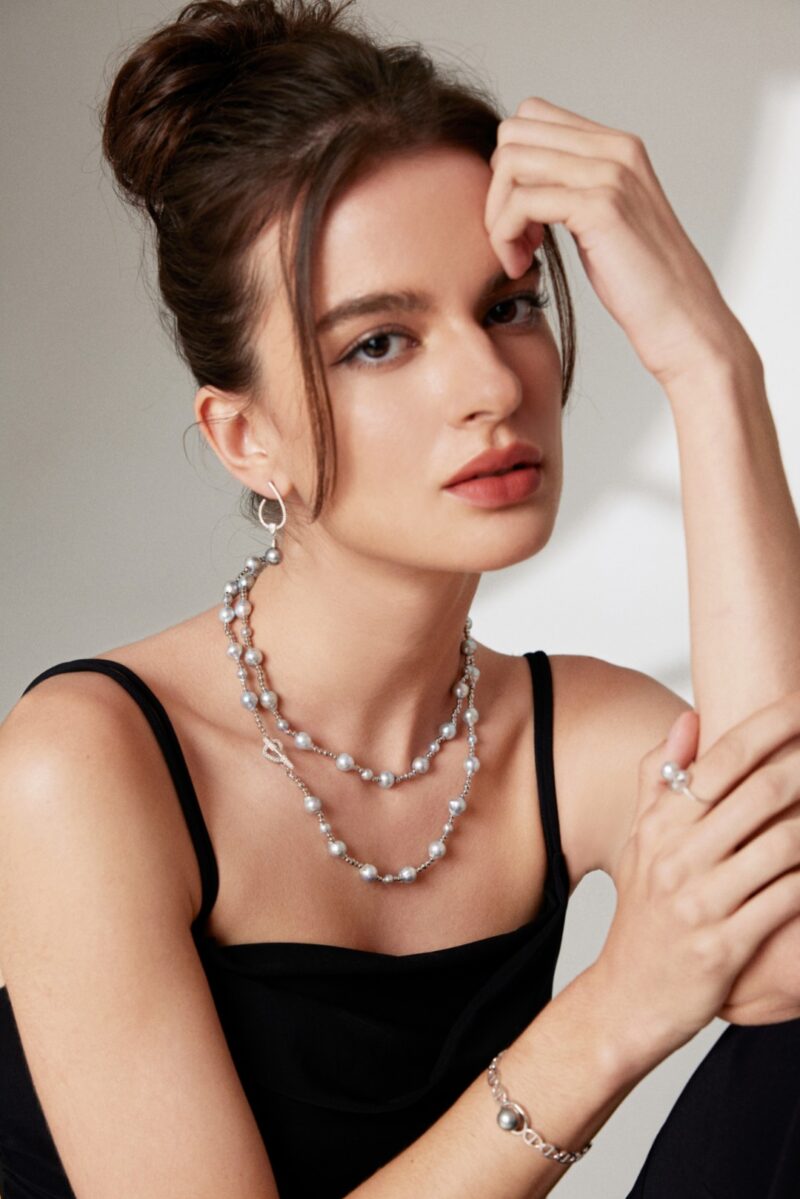 A woman with a thoughtful expression, wearing a black dress and adorned with necklace and large hoop earrings, rests her chin on her hand. Elegant pearl jewelry