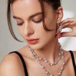 A woman with brown hair and subtle makeup touches her neck gently. She wears a layered necklace and a black top, her eyes closed, emanating a serene expression. Elegant pearl jewelry