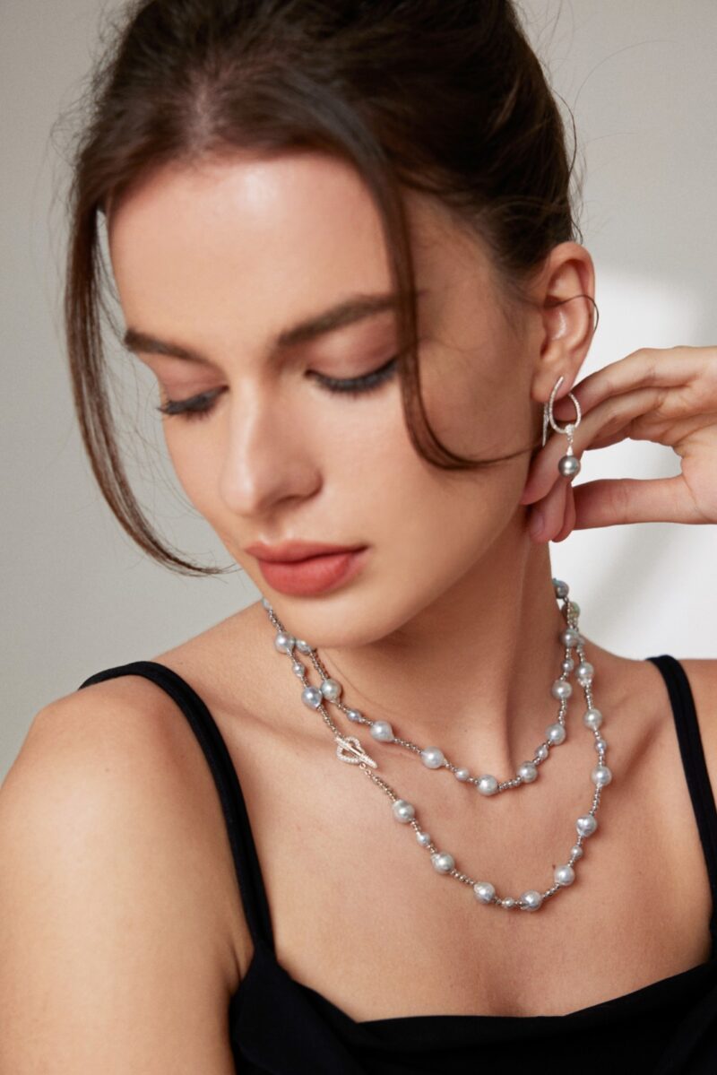 A woman with brown hair and subtle makeup touches her neck gently. She wears a layered necklace and a black top, her eyes closed, emanating a serene expression. Elegant pearl jewelry