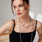 A woman with a bun hairstyle wearing a black sleeveless dress and gold pearl jewelry, looking off to the side thoughtfully, seated against a neutral background. Elegant pearl jewelry