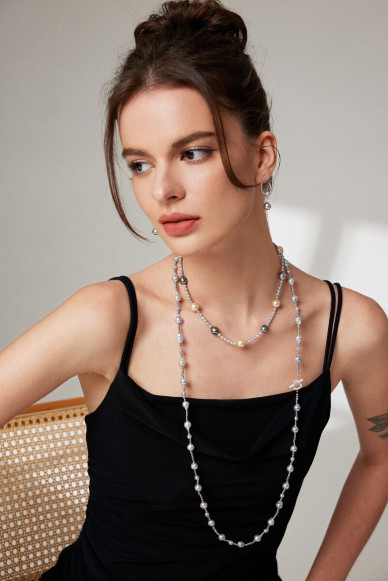 A woman with a bun hairstyle wearing a black sleeveless dress and gold pearl jewelry, looking off to the side thoughtfully, seated against a neutral background. Elegant pearl jewelry