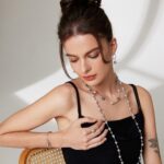 A woman in a black sleeveless top sits thoughtfully, her chin resting on her hand, showcasing a long natural pearl necklace and a small tattoo on her upper arm. Elegant pearl jewelry