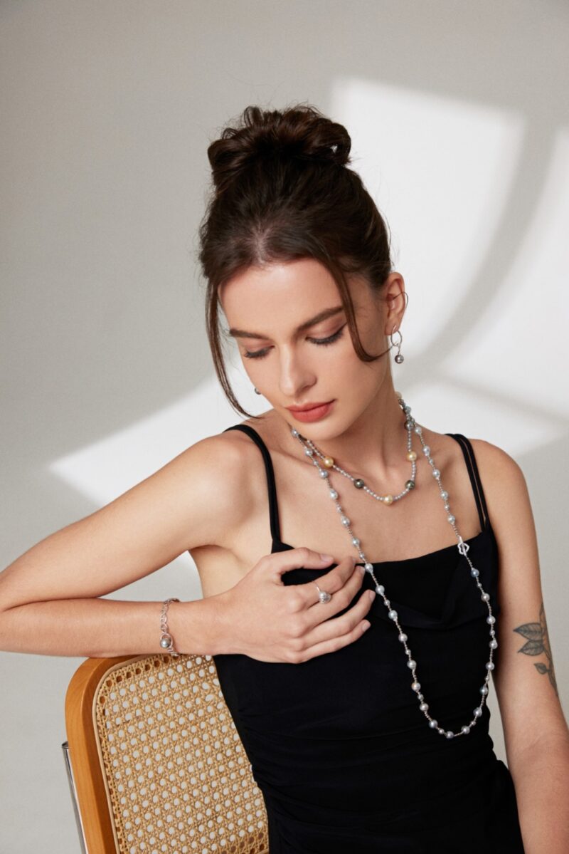 A woman in a black sleeveless top sits thoughtfully, her chin resting on her hand, showcasing a long natural pearl necklace and a small tattoo on her upper arm. Elegant pearl jewelry