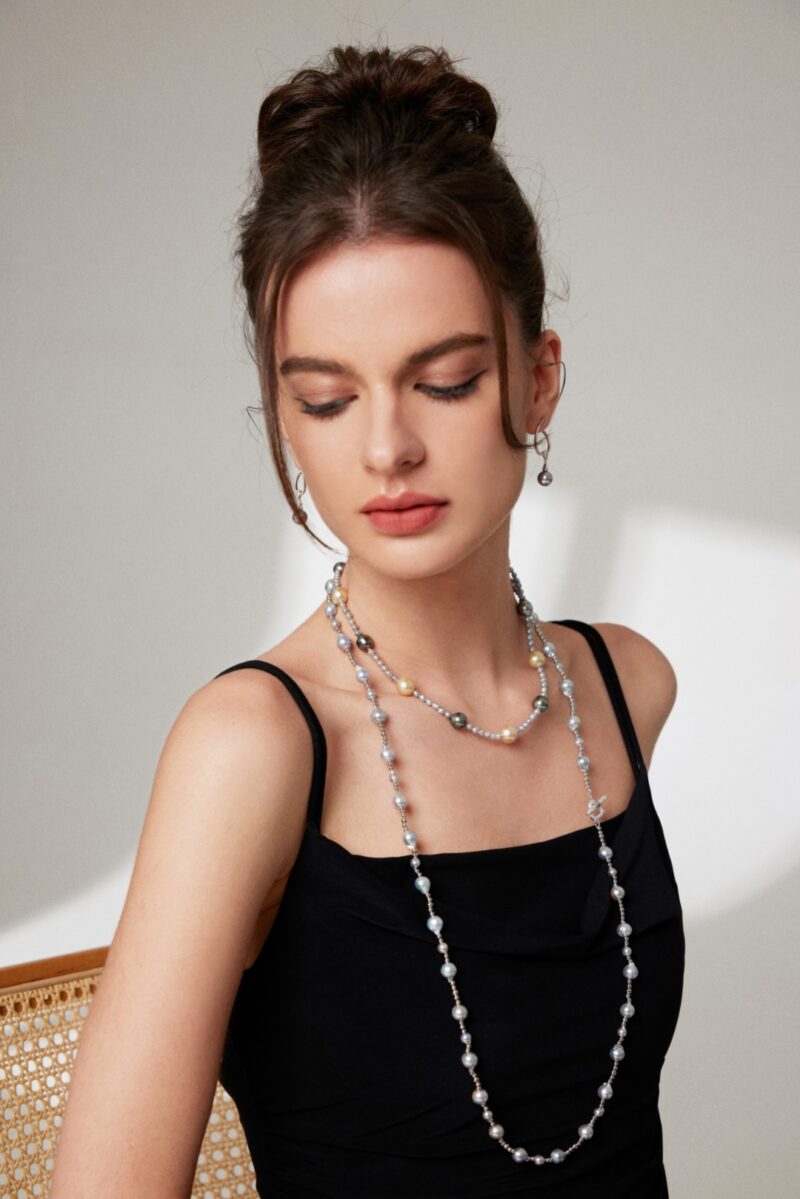 A young woman with a bun hairstyle wearing a black sleeveless top and multiple freshwater pearl necklaces, with closed eyes and a serene expression, sitting indoors, as shown