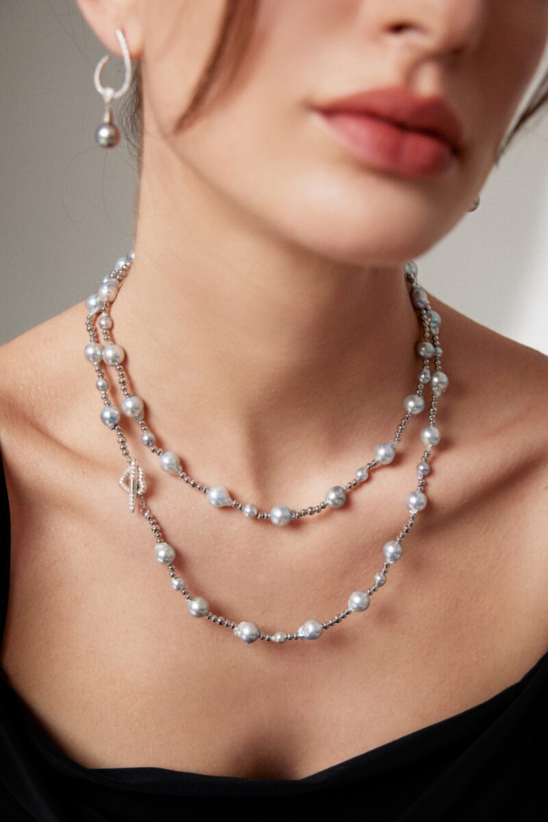 Close-up of a woman wearing a  necklace with a key pendant, and matching earrings, against a soft grey background. Only her neck and lower face are visible. Elegant pearl jewelry