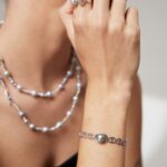 A woman showcases elegant jewelry, featuring a bracelet, a ring, and earrings, set against a blurred background focusing on her adorned hand near her chin. Elegant pearl jewelry