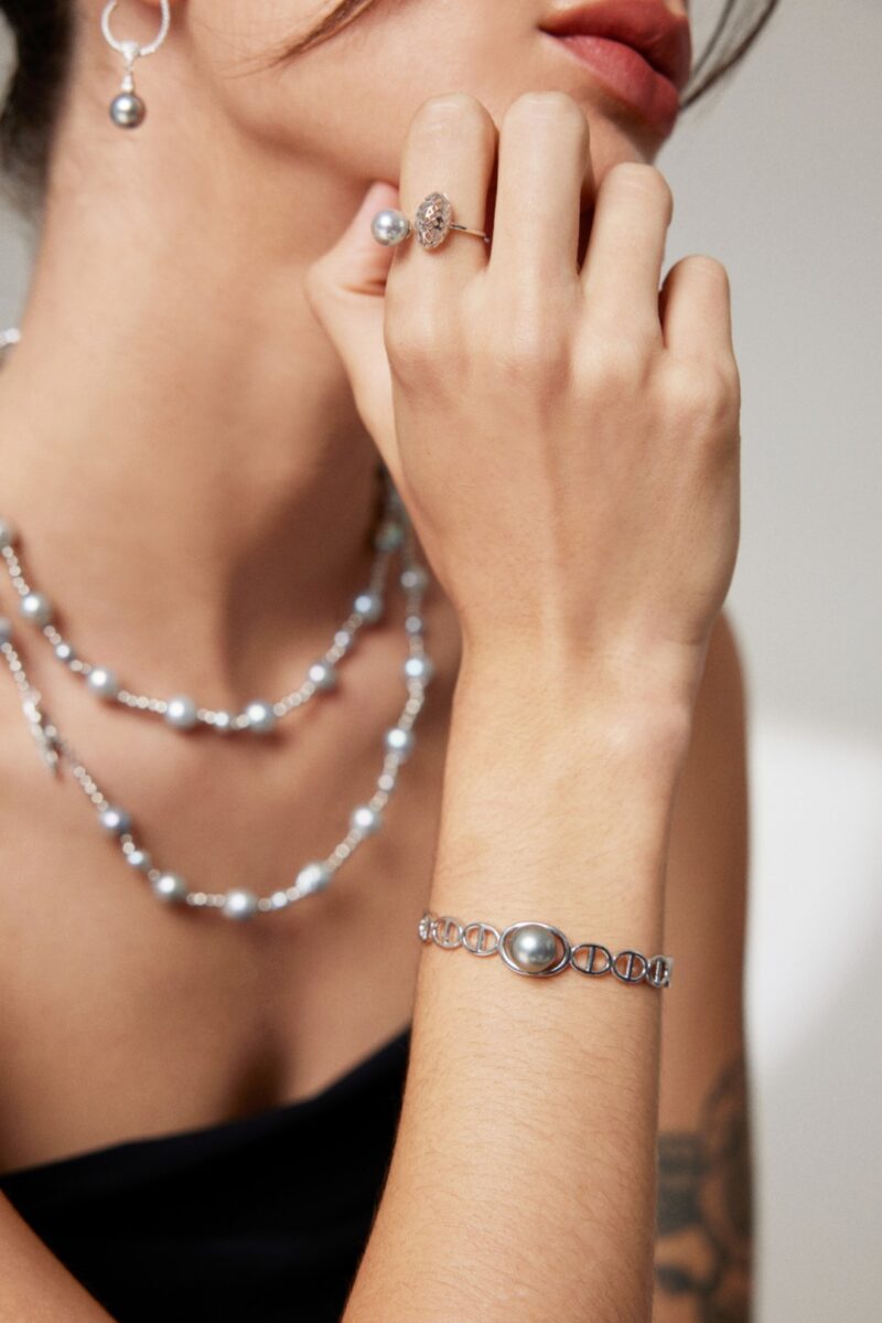 A woman showcases elegant jewelry, featuring a bracelet, a ring, and earrings, set against a blurred background focusing on her adorned hand near her chin. Elegant pearl jewelry