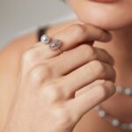 Close-up of a woman's lower face and hand, showcasing a ring and silver necklace, focusing on the elegant details of the jewelry and her pink lipstick. Elegant pearl jewelry