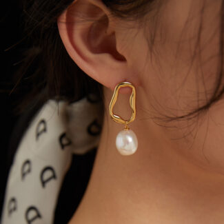 Close-up of a woman's ear wearing a gold earring with the product name design and a dangling freshwater pearl, against a blurred background. Elegant pearl jewelry