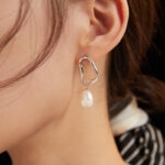 Close-up of a woman's ear wearing unique pearl jewelry, featuring a silver geometric design and a dangling pearl, set against a dark background. Elegant pearl jewelry