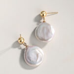 A pair of elegant earrings with a large, iridescent natural pearl dangling from a small gold stud on a light, neutral background. Elegant pearl jewelry