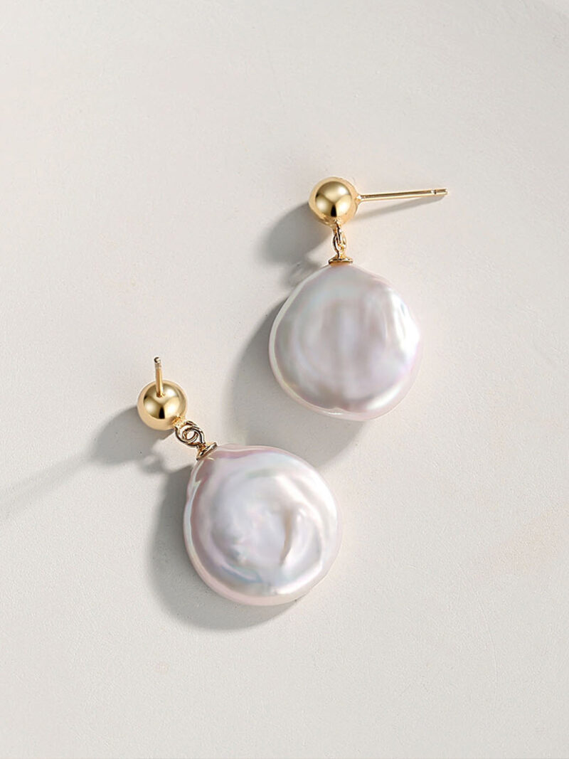 A pair of elegant earrings with a large, iridescent natural pearl dangling from a small gold stud on a light, neutral background. Elegant pearl jewelry