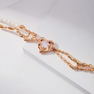 A delicate gold bracelet with a translucent heart-shaped gemstone, displayed on a white geometric background. The bracelet features intricate chain links and natural pearl accents. Elegant pearl jewelry