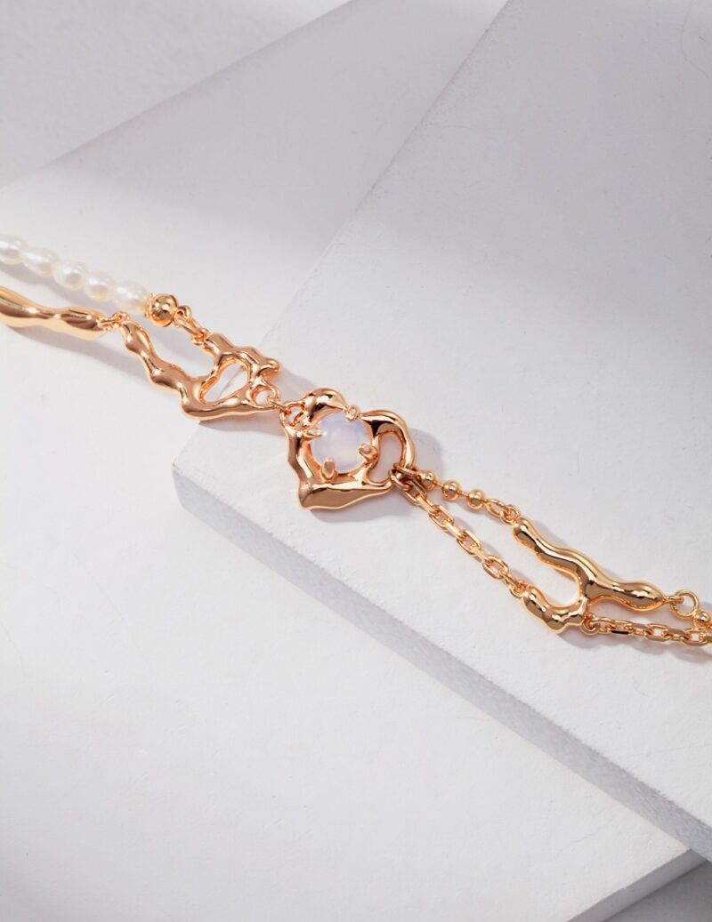 A delicate gold bracelet with a translucent heart-shaped gemstone, displayed on a white geometric background. The bracelet features intricate chain links and natural pearl accents. Elegant pearl jewelry